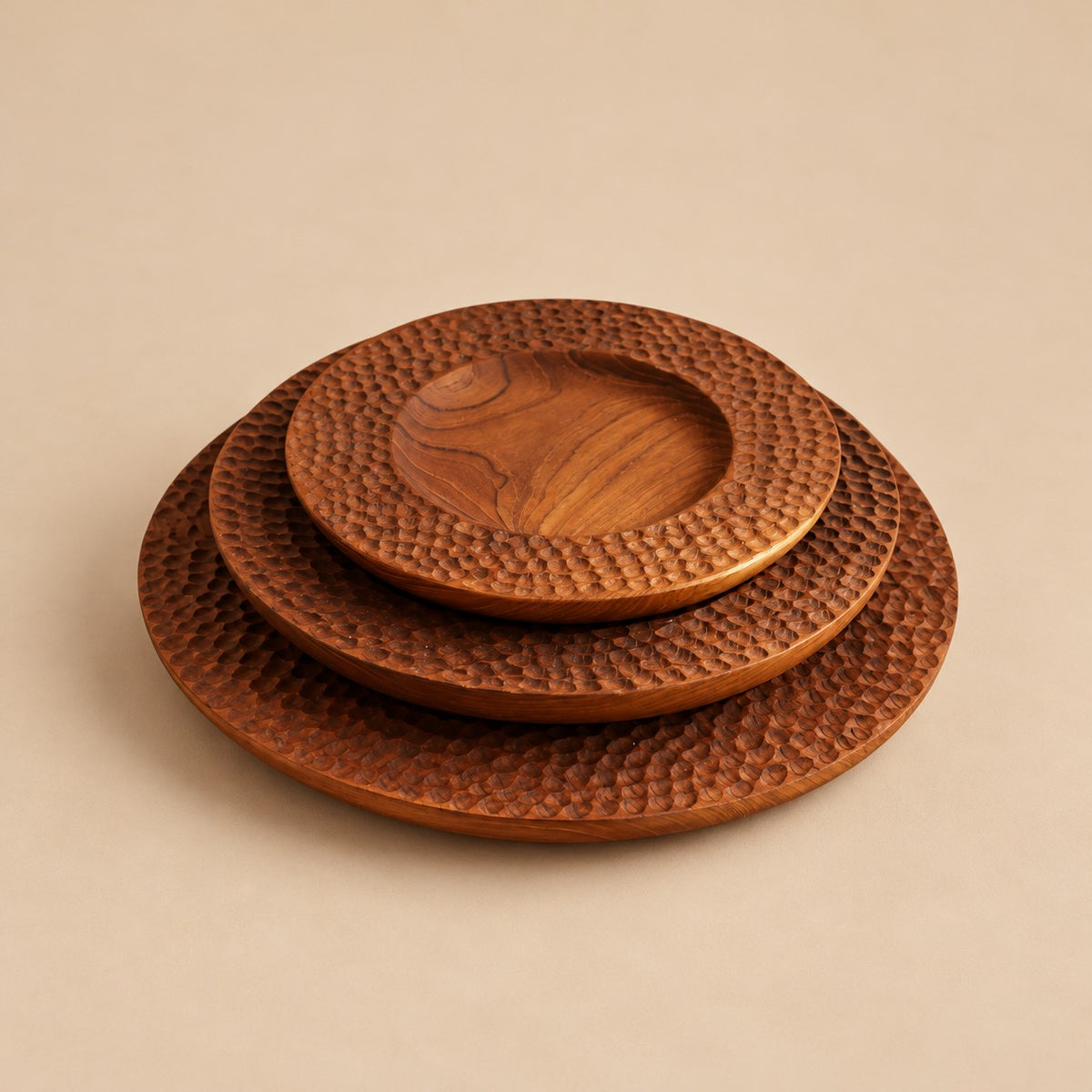 Trois assiettes rondes Carving en teck sculpté empilées sur fond beige, avec bordures texturées.